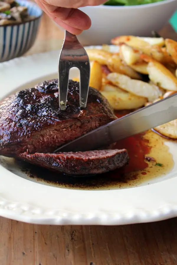 Acción de un cuchillo cortando una carne al horno servida en un plato a un lado de unas papas. 