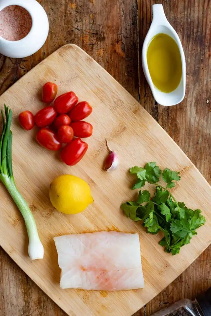 ingredintes sobre una tabla de picar, un filete de bacalo, jitomates cherry, limón eureka, un diente de ajo, una cebolla de cambray, cilantro, aceite de oliva, sal y pimienta.